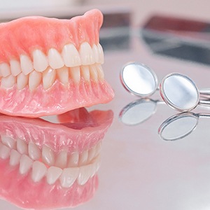 Complete set of dentures on shiny, reflective surface next to 2 dental mirrors