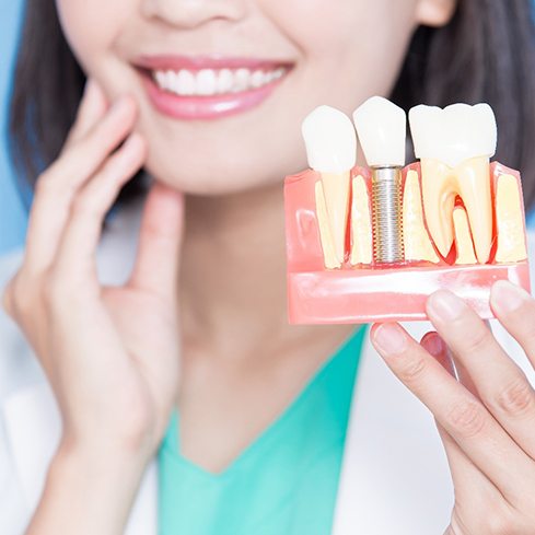 Dentist holding model implant in one hand and touching her cheek with the other