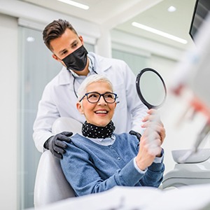 Woman with short white hair in dental chair smiling into mirror with dentist touching her shoulders